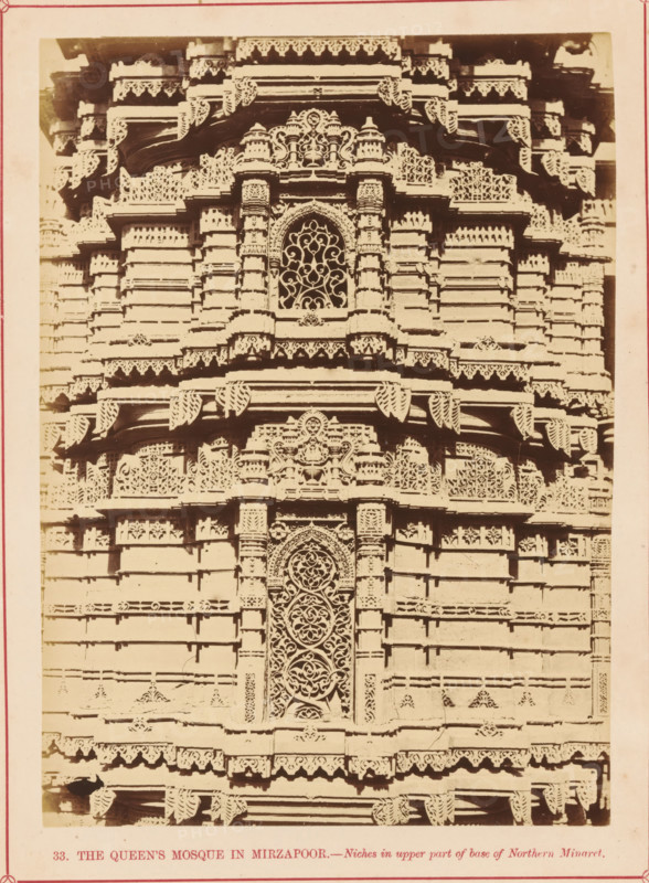 Decoration on the base of a minaret of the Rani Rupamati Mosque in Ahmedabad, The Queen's Mosque in Mirzapoor.- Niches in upper part of base of Northern Minaret, photograph, page, number: '33.', inscription: 'K', photographer: Thomas Biggs, Ahmedabad (city), c. 1856 - in or before 1866, Thomas Biggs, photographer, Gujarat, paper, albumen print, height 188 mm x width 136 mm, temple, shrine, Islam, Mohammedanism, base (, column, pillar), ornamenting, decorating, applied arts, Rani Rupamati Mosque, A close view of an ornate stone façade with layered horizontal bands and carved decorative trim, featuring an arched central niche with an intricate lattice pattern, flanked by carved pilasters and small domed motifs, repeated ornamental brackets and scalloped edges along projecting ledges, and repeated geometric and floral relief panels across the surface.