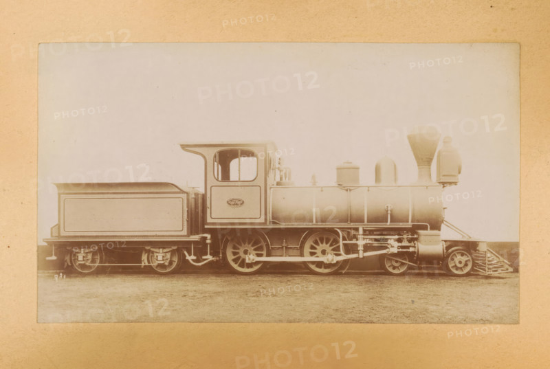 Passenger steam locomotive made by Sharp, Stewart and Company, photograph, page, inscription: 'Class 981. Bogie passenger engine.', number: '981', c. 1874 - in or before 1884, photographer, paper, albumen print, height 83 mm x width 142 mm, steam locomotive, A sepia-toned photograph of a steam locomotive shown in profile, centered on a flat plain, with a rectangular tender behind the cab and a cylindrical boiler toward the front; the cab has an open window and a curved roof edge, the boiler features dome-shaped fittings and a tall funnel, and the front has a cowcatcher and a round headlamp assembly; visible are the large spoked driving wheels, connecting rods, and various mechanical linkages beneath the boiler, with the entire scene framed by a lighter matte border.