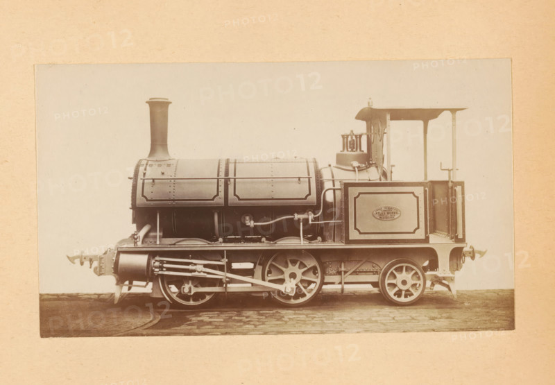 Steam locomotive made by Sharp, Stewart and Company, photograph, page, number: '751', inscription: 'Class 751. Tank engine, for Mixed Traffic.', c. 1874 - in or before 1884, photographer, paper, albumen print, height 81 mm x width 141 mm, steam locomotive, A sepia-toned studio photograph of a vintage steam locomotive shown in profile, with a tall straight chimney rising from a cylindrical boiler that has riveted panels and decorative framed edges, an exposed set of driving wheels with connecting rods and visible mechanical linkages beneath the boiler, a small open cab with a canopy roof and control fittings including valves and pipes, a rectangular side panel on the cab with a framed border, buffer fixtures at the front and rear, and the locomotive positioned on a short section of track against a plain light background with a textured mounting border around the image.
