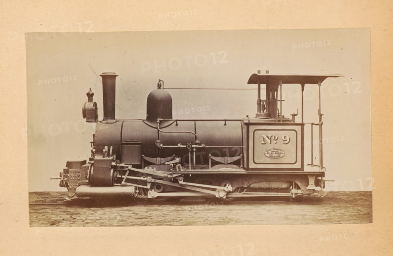 Steam locomotive made by Sharp, Stewart and Company, photograph, page, number: '810', inscription: 'Class 810. Tank engine, for light 2ft. Oin. Gauge Railways.', c. 1874 - in or before 1884, photographer, paper, albumen print, height 83 mm x width 144 mm, steam locomotive, A sepia-toned photograph shows a side view of a steam locomotive mounted on a light background with a textured border, featuring a cylindrical boiler, a tall smokestack near the front, a rounded dome toward the middle, and an open cab area at the rear with a roof supported by slender posts. The front of the locomotive displays a boxy assembly with riveted plates and a small lantern mounted near the smokestack, while the running gear below includes visible pistons, connecting rods, springs, and wheel components. Pipes and handrails run along the side of the boiler, and a rectangular panel is set into the rear side of the locomotive. The ground beneath the locomotive shows cobblestone paving.