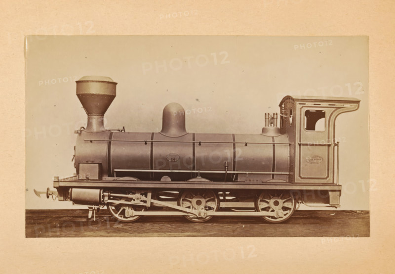 Goods steam locomotive made by Sharp, Stewart and Company, photograph, page, inscription: 'Class 677. Goods engine.', number: '677', c. 1874 - in or before 1884, photographer, paper, albumen print, height 83 mm x width 141 mm, steam locomotive, A sepia-toned photograph of a steam locomotive shown in profile, with a rounded dome on the boiler and a tall, flared smokestack toward the front, mounted on a frame with visible driving wheels and connecting rods; the cylindrical boiler has horizontal bands and handrails, and a rectangular cab at the rear with a curved roof and an open window; beneath the boiler there are cylinders and mechanical linkages, and the whole engine sits on a flat surface against a plain, light background.