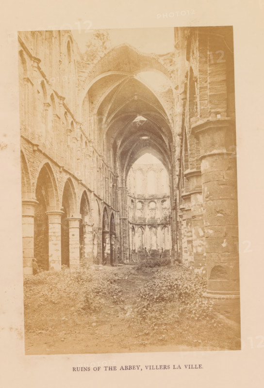 Ruins of the abbey, Villers la ville, photograph, page, Villers-la-Ville, c. 1856 - in or before 1866, photographer, paper, albumen print, height 177 mm x width 118 mm, ruin of church, monastery, Villers Abbey, The image shows a faded, sepia-toned interior view of a large ruined stone building with tall vaulted arches and columns lining both sides, a long central nave leading toward a distant apse with multiple arched openings, broken roof sections revealing sky, rough stone surfaces with visible weathering and chipped areas, overgrown vegetation covering the floor, and scattered rubble along the ground.