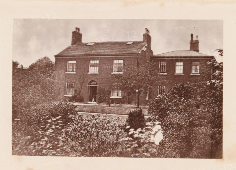 Exterior of John Bright's birthplace at Rochdale, page, photomechanical print, Rochdale, c. 1873 - in or before 1878, photographer, printmaker, Manchester, paper, Woodburytype, height 77 mm x width 118 mm, housing, façade (of house or building), A sepia-toned photograph of a two-story brick house with a tiled roof and multiple chimneys, framed by dense shrubs and trees in the foreground and sides. The house shows several sash windows with visible panes on both floors, an arched central doorway with a closed door, and climbing plants against the facade. A path or driveway runs in front of the house, and trimmed hedges and garden plants occupy the foreground. The sky above the house appears cloudy.