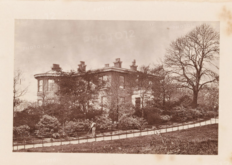 Exterior of (presumably) John Bright's house at Rochdale, page, photomechanical print, Rochdale, c. 1873 - in or before 1878, photographer, printmaker, Manchester, paper, Woodburytype, height 77 mm x width 118 mm, housing, façade (of house or building), A sepia-toned photograph shows a large multi-story brick house partially obscured by dense shrubbery and leafless trees, with a rounded bay window visible at one corner and several chimney stacks rising from the roof; a paved path with a low metal railing runs across the foreground along a grassy slope, and a person wearing light clothing is walking on the path while holding a long tool; a tall bare tree stands to the right of the house and the sky above is pale and empty.