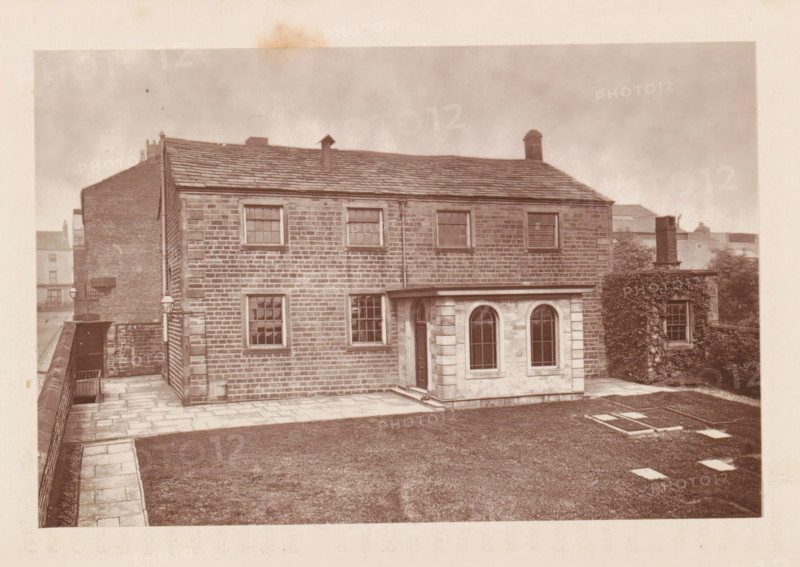Exterior of a house at Rochdale, page, photomechanical print, Rochdale, c. 1873 - in or before 1878, photographer, printmaker, Manchester, paper, Woodburytype, height 77 mm x width 118 mm, housing, façade (of house or building), A stone building with a pitched slate roof fills the frame, showing a rectangular facade of evenly coursed masonry with several sash windows and a downpipe running vertically; a small single-story projection with arched windows and a doorway extends from the front, and a paved path and low stone wall border a lawn area in the foreground, while an adjacent wall covered in climbing plants and a chimney are visible to one side and rooftops of other buildings appear in the background.