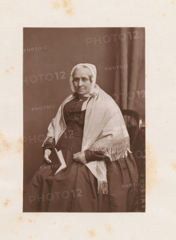Studio portrait of Hannah S. Allen, page, photomechanical print, c. 1874 - in or before 1884, photographer, printmaker, paper, Woodburytype, height 90 mm x width 59 mm, Print on frontispiece., Person, Hannah S. Allen, historical persons - BB - woman, A seated older woman wearing a dark dress and a light shawl with fringe is shown from head to mid skirt, her hair covered by a lace-trimmed bonnet, holding a closed book in one hand while the other rests on her lap, a dark chair back is visible behind her and a plain curtain hangs to one side, the photograph is bordered by a wide light-colored mount with scattered small stains.