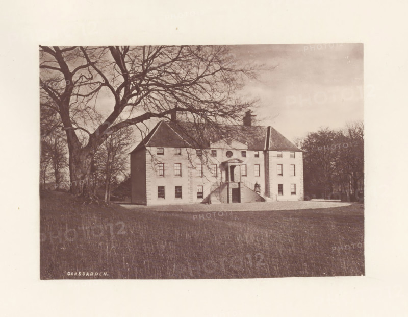 Garscadden, photograph, page, photographer: Thomas Annan, Glasgow, c. 1868 - in or before 1878, paper, carbon print, height 115 mm x width 160 mm, rural housing, eg country-house, villa, cottage, façade (or house or building), A large pale building with a central staircase and a portico is shown set on a grassy foreground, bare-branched trees frame the scene with a very large tree on the left whose branches extend across the sky, the building has multiple windows and a pitched roof with a chimney, a person stands on the staircase near the entrance, and trees and shrubbery are visible behind and to the sides of the building.