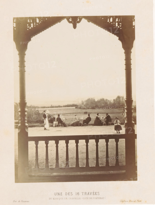 Company in front of the kiosk on the Plateau de Gravelle, Une des 16 travées du kiosque de Gravelle (côté de Pontenay), Bois de Vincennes, photograph, page, photographer: Ildefonse Rousset, Plateau de Gravelle, c. 1861 - in or before 1866, Paris, paper, albumen print, height 163 mm x width 123 mm, wood and park (as places of recreation), summer-house, arbour, garden-house (Dutch: 'tea dome'), A sepia photograph framed by ornate wooden posts and a carved roof edge shows a group of people seated at outdoor tables on a lawn, with a low fence and distant trees in the background; several seated figures wear long coats or dresses and hats, one standing figure in a uniform or apron walks near the group, and a small child stands closer to the foreground; a wooden balustrade fills the lower foreground, and the scene is lit with soft, even daylight.