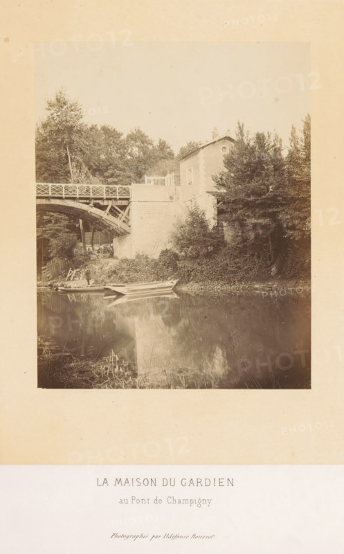 Bridge keeper's house of the Pont de Champigny in Champigny-sur-Marne, La maison du gardien au Pont de Champigny, photograph, page, photographer: Ildefonse Rousset, Île-de-France, c. 1860 - in or before 1865, Ildefonse Rousset, photographer, paper, albumen print, height 152 mm x width 122 mm, river, bridge, housing, Marne (river), Pont de Champigny, Champigny-sur-Marne, A sepia-toned photograph shows a riverbank scene with a wooden arched bridge spanning from the left toward a stone embankment, a small house partially hidden by trees perched above the embankment, dense foliage and trees surrounding the house and bridge, a narrow boat moored at the water’s edge near a small grassy and rocky slope, the water reflecting the boat, embankment, and nearby vegetation, and a wide pale margin around the photograph with a printed caption area beneath the image.