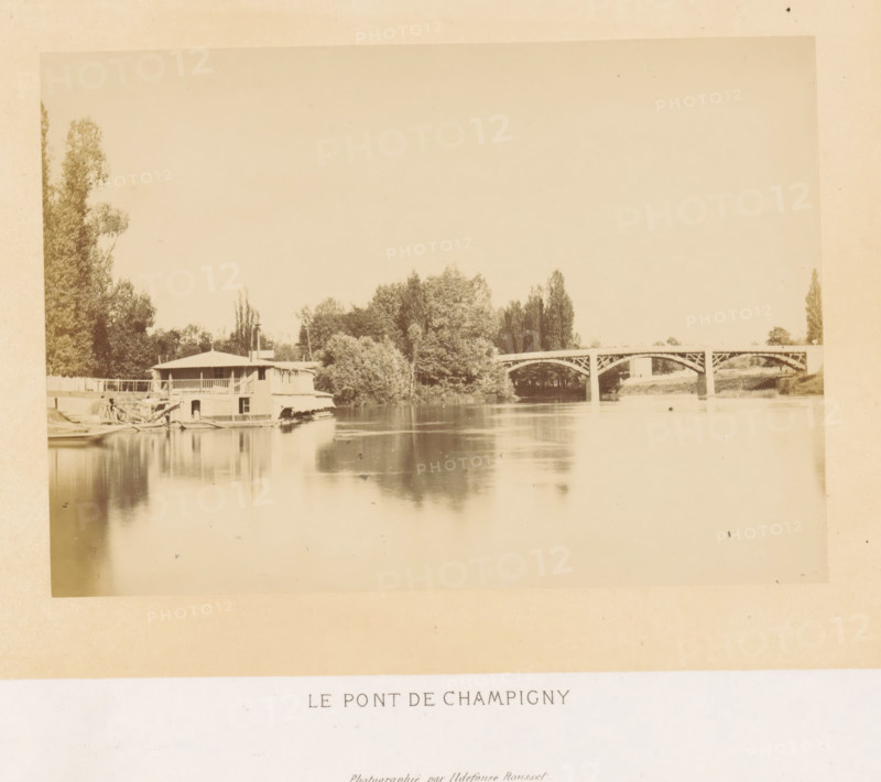 View of the Pont de Champigny in Champigny-sur-Marne, Le pont de Champigny, photograph, page, photographer: Ildefonse Rousset, Île-de-France, c. 1860 - in or before 1865, paper, albumen print, height 123 mm x width 178 mm, river, bridge, Marne (river), Pont de Champigny, Champigny-sur-Marne, A sepia-toned riverscape showing a calm stretch of water reflecting the sky and surrounding features, with a low two-story riverside building with a covered balcony and small boats moored beside it at the near left, dense clusters of trees rising behind and along the riverbank toward the center, and an arched bridge with decorative supports crossing the river toward the right, with more trees and grassy banks visible beyond the bridge.