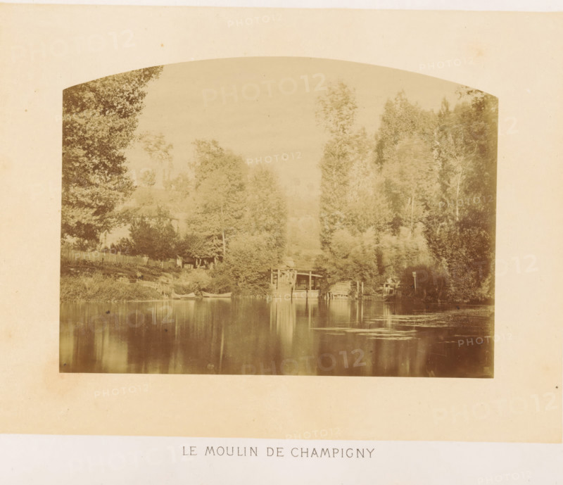 Le moulin de Champigny, Marne in Champigny-sur-Marne, photograph, page, photographer: Ildefonse Rousset, Champigny-sur-Marne, c. 1860 - in or before 1865, paper, albumen print, height 115 mm x width 156 mm, river, Marne (river), A faded sepia photograph shows a riverside scene framed by tall trees on both sides, their foliage dense and varied in texture. Along the near bank there is a wooden fence and a small building partially obscured by trees. A wooden structure extends over the water, with a dock area and what appear to be small boats moored nearby. The water surface is calm and reflective, mirroring the trees and structures with soft, vertical ripples and patches of lighter texture. The overall image has an arched top and a broad pale border surrounding it.