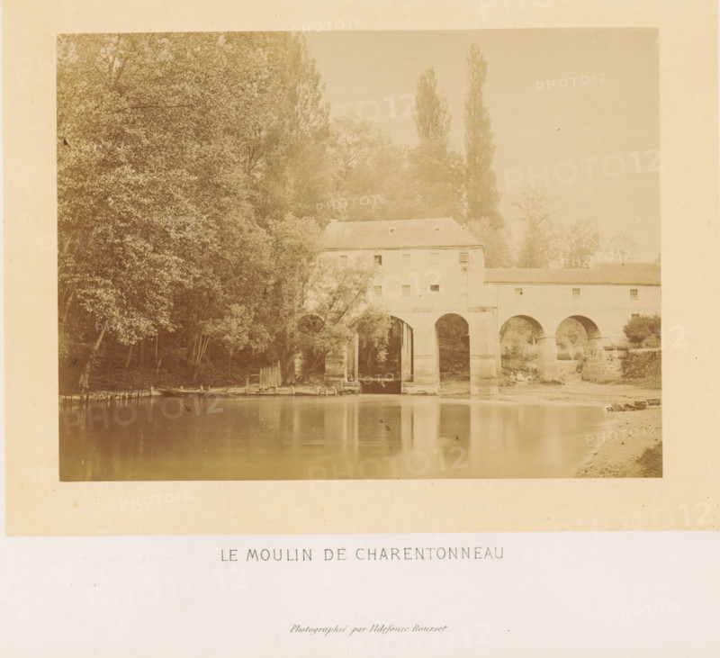 Le moulin de Charentonneau, Exterior of the Moulin Brûlé in the Marne, photograph, page, photographer: Ildefonse Rousset, Île-de-France, c. 1860 - in or before 1865, paper, albumen print, height 128 mm x width 173 mm, river, watermill, façade (of house or building), Marne (river), A sepia-toned photograph showing a riverside scene with a stone building spanning the water on large arched supports, trees and foliage clustered to the left and behind the building, reflections of the arches and shoreline visible in the calm river, a small wooden dock and a few small boats along the near bank, and a narrow strip of sandy shore on the right.
