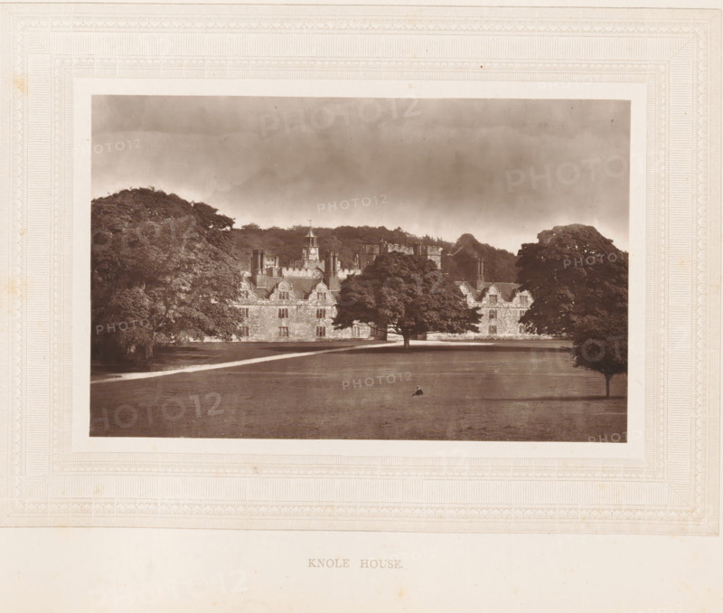 Knole House, View of Knole House, page, photomechanical print, photographer: Stephen Thompson, Kent, c. 1866 - in or before 1876, printmaker, paper, Woodburytype, height 102 mm x width 160 mm, country-house, A sepia-toned photograph framed by an ornate embossed border shows a large stone building with multiple chimneys and gabled rooflines partially obscured by mature trees; a wide expanse of lawn stretches in front of the building with a single small figure seated near the center, and a gravel path runs parallel to the facade beneath a cloudy sky.