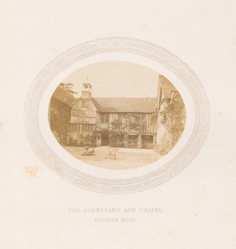 Courtyard and chapel of Ightham Mote near Ightham, The courtyard and chapel, Ightham Mote, photograph, page, photographer: Stephen Thompson, Ightham, c. 1866 - in or before 1876, Kent, paper, albumen print, albumendruk, height 70 mm x width 89 mm, courtyard, rural housing, eg country-house, villa, cottage, church (exterior) - QQ - small church, chapel, A sepia-toned circular photograph framed by an ornate patterned border shows a timber-framed building with a pitched roof and a small bell tower above one gable; the facade features exposed beams, mullioned windows, and an open arcade at ground level partially covered in climbing plants, with a gravel courtyard in front containing a decorative stone urn on a pedestal and a folded metal chair, flanked by trees and foliage on either side.