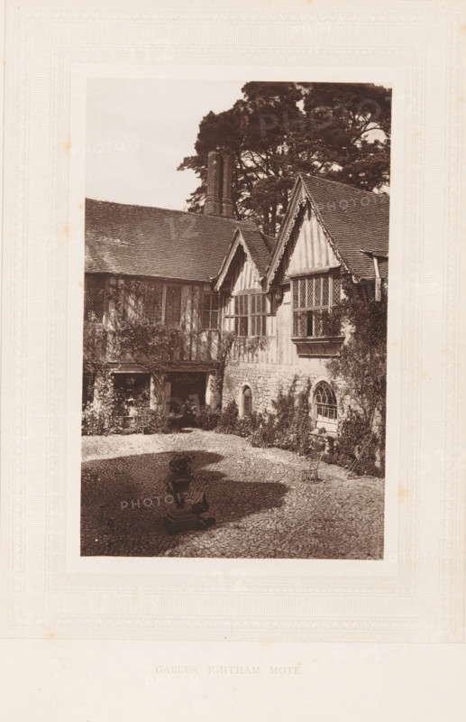 Gables, Ightham Mote, View of the Ightham Mote near Ightham, page, photomechanical print, photographer: Stephen Thompson, Ightham, c. 1866 - in or before 1876, printmaker, Kent, paper, Woodburytype, height 160 mm x width 102 mm, courtyard, rural housing, eg country-house, villa, cottage, A sepia-toned photograph shows a courtyard framed by a timber-framed building with steep gabled roofs and leaded lattice windows, climbing plants on the walls, and a stone lower façade with an arched window; a tall chimney rises behind the roofline and large trees fill the background; the cobbled courtyard contains a decorative urn on a plinth casting a shadow and a small metal chair near the building, with a shaded recess under the eaves where foliage and a small table are partially visible.