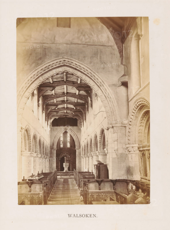 Walsoken, photograph, page, photographer: Edward Johnson, All Saints' Church (Walsoken), c. 1858 - in or before 1869, Edward Johnson, photographer, Cambridgeshire, paper, albumen print, height 178 mm x width 126 mm, interior of church, facade (of house or building), A sepia photograph showing the interior of a church nave with a central aisle flanked by wooden pews and carved choir stalls, a patterned stone floor leading toward a raised pulpit or font in the distance, tall arched openings and columned arcades along the side walls, ornate stone archways with decorative moldings, a timber roof with exposed beams and trusses, high walls with plastered surfaces, stained glass windows visible at the far end, and various stone and wooden architectural details and furnishings along the sides.
