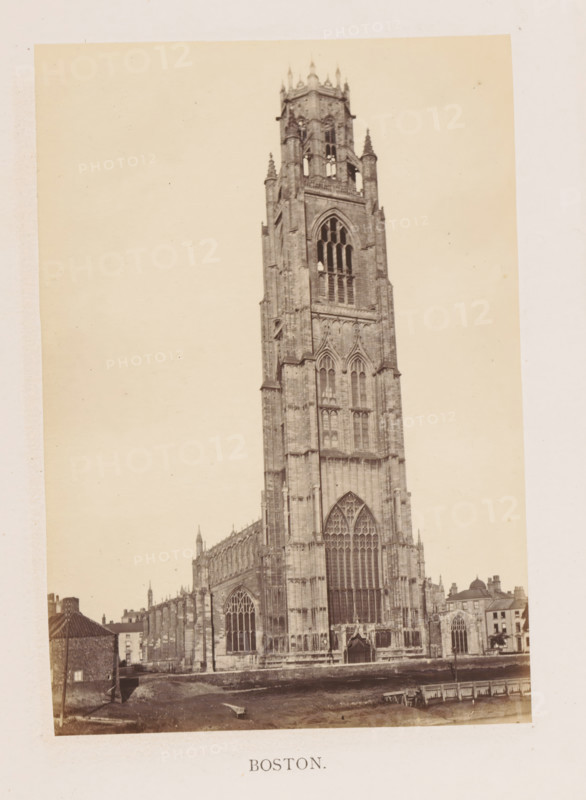 Boston, photograph, page, c. 1858 - in or before 1869, photographer, Lincolnshire, paper, albumen print, height 175 mm x width 122 mm, church (exterior), facade (of house or building), Saint Botolph's Church, A sepia-toned photograph of a tall Gothic stone cathedral tower with ornate pointed arches, large stained-glass window openings, carved stone tracery, buttresses, and pinnacles, flanked by the lower nave and aisles with rows of arched windows, adjacent smaller buildings with pitched roofs, a foreground area of bare ground and a low retaining wall, and a pale blank sky above.