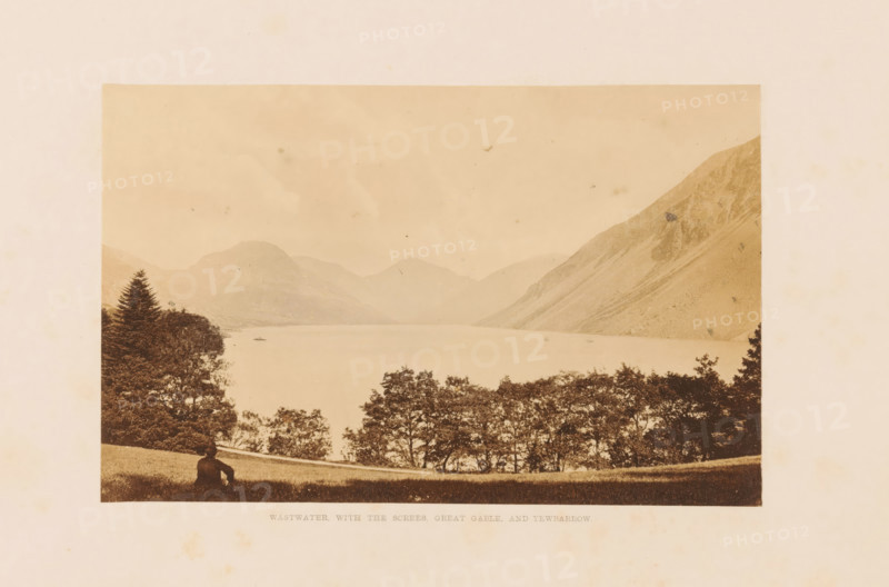 View of Wast Water and mountains, Wastwater, with the screes, Great Gable, and Yewbarrow, photograph, page, photographer: Garnett & Sproat (possibly), Lake District, c. 1857 - in or before 1871, paper, albumen print, height 100 mm x width 158 mm, lake, mountains, Wast Water, A sepia-toned landscape photograph showing a wide lake bordered by tree-covered shores and steep mountains in the background, with a line of trees along the near shoreline and grassy foreground where a solitary person sits facing the water, a tall conifer rising on the left and rugged slopes descending into the lake on the right, all framed by a pale border.