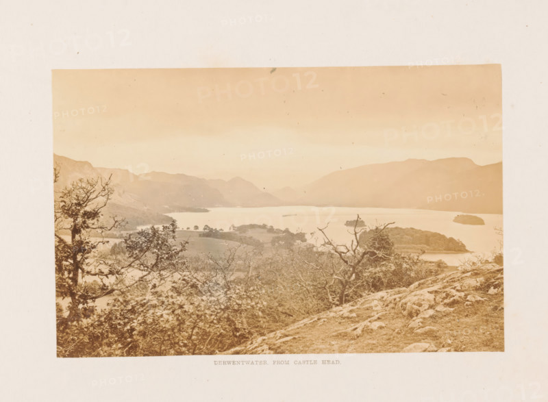 View of Derwentwater in the Lake District, Derwentwater, from Castle head, photograph, page, photographer: Garnett & Sproat (possibly), Lake District, c. 1857 - in or before 1871, paper, albumen print, height 101 mm x width 157 mm, lake, mountains, A wide sepia-toned landscape photograph showing a lake surrounded by rolling hills and distant mountains under a light sky, with islands and shoreline visible on the water, trees and bare-branched shrubs in the foreground growing on rocky, grassy terrain, and scattered small dark marks and spots across the photograph.