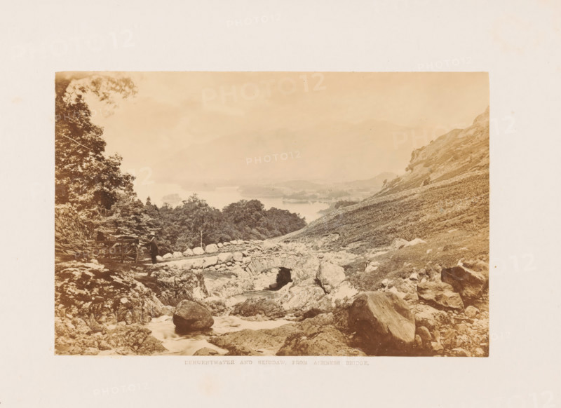 Derwentwater and Skiddaw, from Ashness Bridge, View of Derwentwater and Mount Skiddaw in the Lake District, photograph, page, photographer: Garnett & Sproat (possibly), Lake District, c. 1857 - in or before 1871, paper, albumen print, height 107 mm x width 164 mm, lake, mountains, Derwentwater, Skiddaw, A sepia-toned landscape photograph showing a rocky hillside and stream in the foreground with a stone arch bridge spanning the water and scattered boulders and fallen logs nearby, a person wearing a hat standing beside a wooden cart or gate near a rocky path, dense trees and shrubs on the left, a sloping grassy hillside rising on the right, and a distant view of misty hills and a large body of water beyond the tree line under a pale sky.