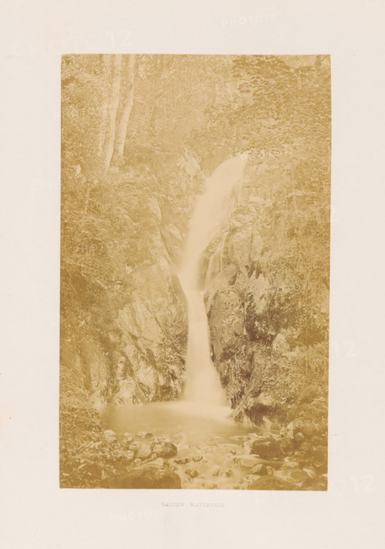 Barrow Waterfall, View of a waterfall in the Lake District, photograph, page, photographer: RJ Sproat, Lake District, c. 1857 - in or before 1870, paper, albumen print, height 166 mm x width 103 mm, waterfall, A tall, narrow sepia-toned photograph of a waterfall framed by steep rocky cliffs and dense foliage, showing water cascading down a narrow chute into a calm pool surrounded by scattered rocks, with trees and undergrowth visible along the cliff edges and rocky surfaces textured with moss and crevices.