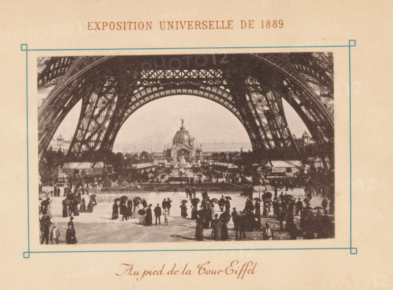 La Tour Eiffel, View of the Eiffel Tower in Paris, Exposition Universelle de 1889, page, photomechanical print, maker: Neurdein Frères, Paris, 1889, paper, collotype, photoprint, height 108 mm x width 69 mm, tower, clock tower, monumental adornment, world fair, world exhibition, World Exhibition 1889 (Paris), Eiffel tower, A sepia-toned photograph framed by a thin blue border shows the underside of a large iron lattice structure forming a sweeping arch above a crowded open space, with ornate metalwork visible along the arch and intricate cross-bracing in the supporting legs; beneath the arch a domed pavilion with sculptural details and fountains sits at the center of a landscaped area, flanked by trees and additional architectural elements; in the foreground a crowd of people dressed in period clothing, many holding umbrellas and wearing long coats and hats, are walking and standing on a paved area, with smaller figures and grouped clusters extending toward the pavilion and the edges o...