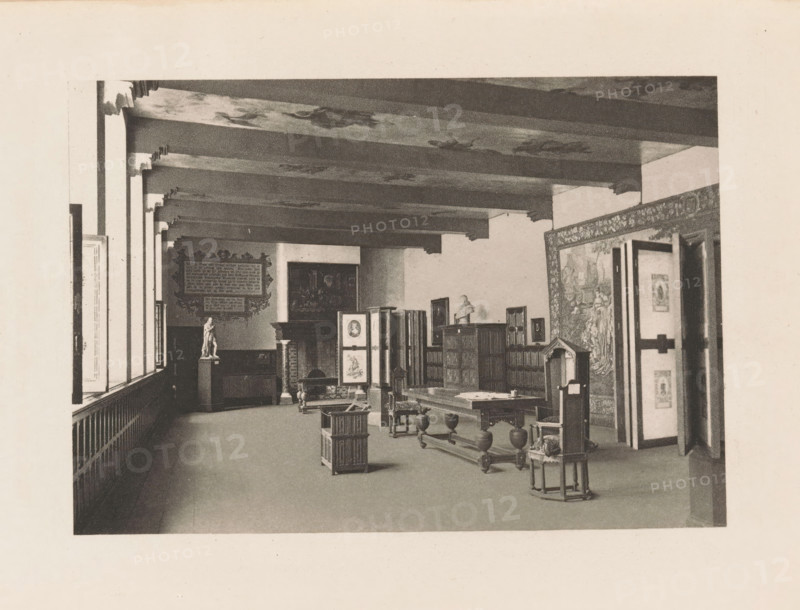 Refectory of the former St. Agatha Monastery in Delft, page, photomechanical print, title: 'Refectory of the Convent of St. Agatha, Delft.', photographer: Charles L. Mitchell, Prinsenhof (Delft), cliché maker: AW Elson & Company, Boston (Massachusetts), c. 1884 - in or before 1894, Delft, North America, Massachusetts, paper, photoaquatint, height 87 mm x width 124 mm, interior, representation of a building, refectory, monastery, A long interior room with exposed ceiling beams and patterned ceiling panels, a row of tall windows along the left side, wooden wainscoting beneath the windows, and a decorative framed panel on the far wall. Various pieces of wooden furniture are arranged in the space including tables, chairs, and cabinets, a folding screen with framed images, and a pedestal topped by a small statue. A large tapestry or mural covers the right wall near an open doorway, and smaller framed artworks are displayed on the walls and atop furniture. The floor is open and mostly clear ...