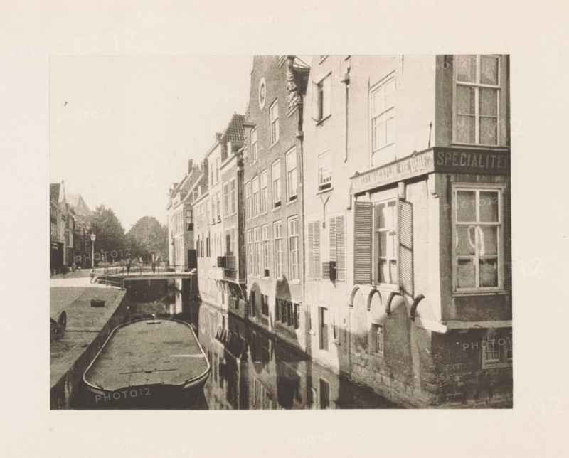 View of a canal in Delft, page, photomechanical print, title: 'Old Delft.', photographer: Charles L. Mitchell, Delft, cliché maker: AW Elson & Company, Boston (Massachusetts), c. 1884 - in or before 1894, North America, Massachusetts, paper, photoaquatint, height 88 mm x width 113 mm, canals, waters (in city), A narrow canal runs alongside a row of tall, narrow buildings with multiple windows and shutters, the canal surface reflecting the building facades; a moored flat boat occupies the foreground with another vessel partially visible; a small arched bridge spans the canal in the midground and a tree-lined street with lampposts appears beyond it; building details include decorative gables, window boxes, and rounded masonry at the waterline, and a large window with shutters and curtains is visible on the right.