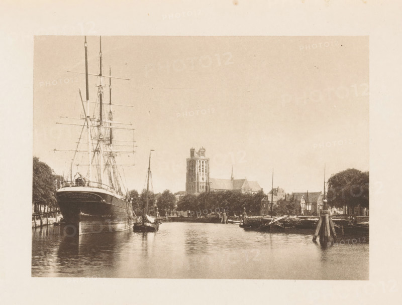 View of the Meuse at Dordrecht, with the Grote or Onze-Lieve-Vrouwekerk in the background, page, photomechanical print, title: 'Dordrecht-Canal with Cathedral in the Distance.', photographer: Charles L. Mitchell, Dordrecht, clichémaker: AW Elson & Company, Boston (Massachusetts), c. 1884 - in or before 1894, North America, Massachusetts, paper, photoaquatint, height 88 mm x width 123 mm, river, church (exterior), ships (in general), Grote or Onze-Lieve-Vrouwekerk, A sepia-toned waterfront scene showing a large ship with tall masts moored at the left edge, its bow facing forward and rigging lines visible, smaller boats alongside and reflections in the calm water; a riverside town stretches across the middle distance with trees along the bank, a prominent multi-story tower and church spire rising above lower buildings, wooden posts and piers extending into the water at right, and an expanse of pale sky above.