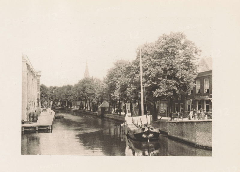 View of a canal in Delft, page, photomechanical print, title: 'Near the Arsenal, Delft.', photographer: Charles L. Mitchell, Delft, cliché maker: AW Elson & Company, Boston (Massachusetts), c. 1884 - in or before 1894, North America, Massachusetts, paper, photoaquatint, height 87 mm x width 117 mm, canals, waters (in city), A sepia-toned scene shows a narrow canal lined with buildings and trees, with a sailboat moored in the foreground reflecting in the water; a row of trees extends along the canal bank, and small boats and houseboats are visible along the water; on the right a quay with a railing borders buildings with windows and awnings, and a few people stand near the railing; on the left a dock area with barrels and steps leads to the canal, and a distant church spire rises above the treeline.