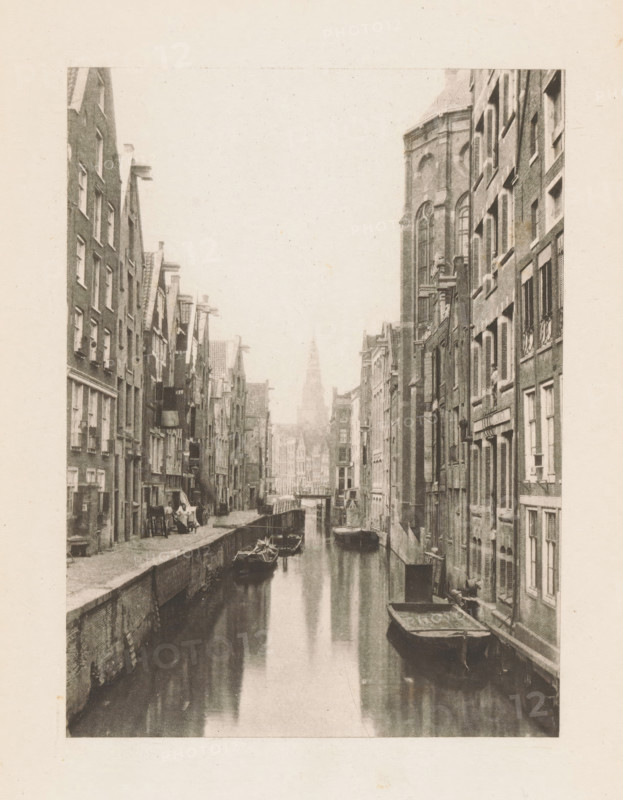 Canal in Amsterdam, page, photomechanical print, title: 'Old Amsterdam.', photographer: Charles L. Mitchell, Amsterdam, cliché maker: AW Elson & Company, Boston (Massachusetts), c. 1884 - in or before 1894, North America, Massachusetts, paper, photoaquatint, height 120 mm x width 88 mm, Print on frontispiece., canals, waters (in city), A narrow canal runs between tall brick buildings on both sides, with moored wooden boats floating on the water and a small bridge visible farther down the channel; the buildings show multiple windows and doors, some with shutters and decorative architectural details, and a larger building with tall arched windows rises prominently on the right; the canal edges have stone walkways and occasional small items and a few indistinct figures near the waterside; the scene is rendered in a sepia-toned, aged photographic style.