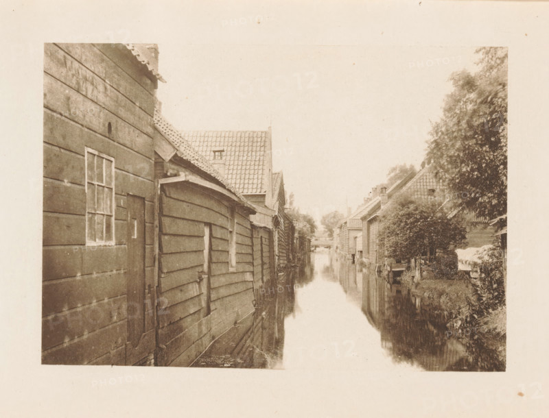 Houses along a ditch in Zaandam, page, photomechanical print, title: 'Afternoon in Zaandam.', photographer: Charles L. Mitchell, Zaandam, cliché maker: AW Elson & Company, Boston (Massachusetts), c. 1884 - in or before 1894, Zaanstreek, North America, Massachusetts, paper, photoaquatint, height 88 mm x width 125 mm, housing, canals, waters (in village), A narrow waterway runs down the center of the image, flanked by wooden buildings with slatted siding on the left and a mix of pitched roof houses and dense foliage on the right, their reflections visible in the water; the left structures show a small window and a door, and the rooftops display overlapping tiles and eaves, while the right side features trees and shrubs near the banks and a covered object near the edge.