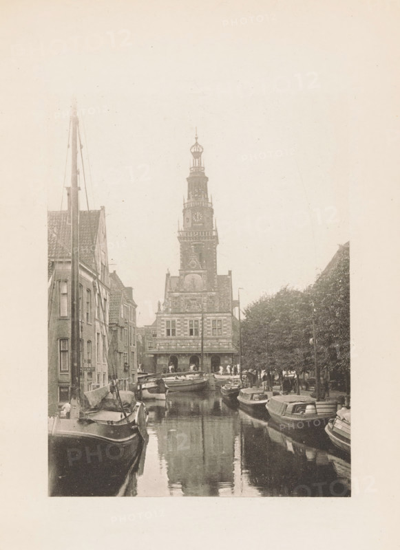 View of the Waag in Alkmaar, page, photomechanical print, title: 'The Town Weighing House, Alkmaar.', photographer: Charles L. Mitchell, Alkmaar, cliché maker: AW Elson & Company, Boston (Massachusetts), c. 1884 - in or before 1894, North America, Massachusetts, paper, photoaquatint, height 118 mm x width 90 mm, canals, waters (in village), façade (of house or building), Waag (Alkmaar), A narrow canal lined with moored boats on both sides leads toward a tall ornate building with a multi-tiered tower and decorative facade; narrow brick buildings flank the left side of the canal while trees and lamp posts line the right side, and the water reflects the boats and the tower above.
