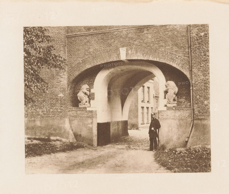 Broederpoort in Kampen, page, photomechanical print, title: 'Archway of Monks' Gate, Kampen.', photographer: Charles L. Mitchell, Kampen, cliché maker: AW Elson & Company, Boston (Massachusetts), c. 1884 - in or before 1894, Charles L. Mitchell, AW Elson & Company, photographer, cliché maker, North America, Massachusetts, paper, photoaquatint, height 87 mm x width 108 mm, gate, fortified city, Broederpoort, A sepia-toned image of a brick archway leading into a courtyard, with two sculpted lion figures perched on short plinths at either side of the arch, the arch showing a rounded inner passage and a rectangular opening beyond where a multiwindowed building is visible, a uniformed man standing near the right side of the passage holding a cane, brick walls flanking the arch with ivy visible on the left wall and a vertical pipe running down the right wall, a dirt pathway and low vegetation in the foreground.
