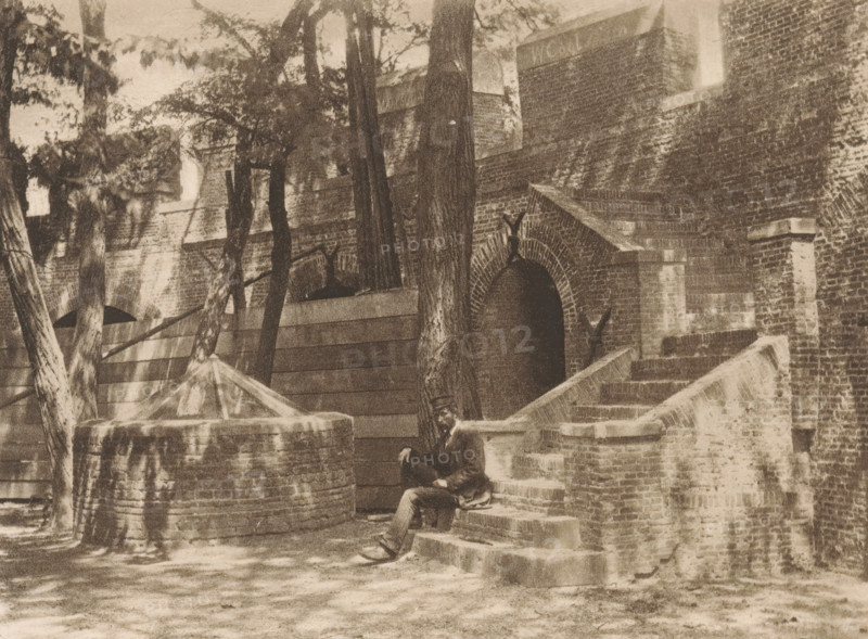 Inside of the Castle of Leiden, page, photomechanical print, title: 'Courtyard of the Burg, Leiden.', photographer: Charles L. Mitchell, Leiden, cliché maker: AW Elson & Company, Boston (Massachusetts), c. 1884 - in or before 1894, North America, Massachusetts, paper, photoaquatint, height 86 mm x width 116 mm, castle, walls, fortified city, Burcht van Leiden, A sepia-toned scene of a brick building with arched entryways and a flight of stone steps leading up to one of the openings, flanked by low brick walls and rounded stone structures; tall trees with textured trunks and leafy branches stand in front of the building, casting dappled shadows across the ground and walls; a man in dark clothing and a hat sits on the steps near the base of a tree, with one leg extended and one hand resting on his knee; the foreground shows a ground surface with scattered light patches and the image is bordered by a pale margin.