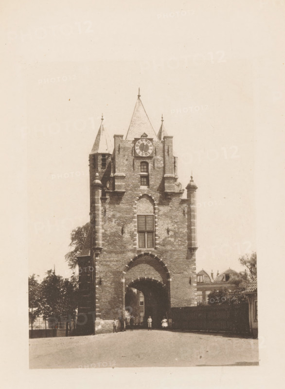 Amsterdam Gate in Haarlem, page, photomechanical print, title: 'The Amsterdam Gate, Haarlem.', photographer: Charles L. Mitchell, Haarlem, cliché maker: AW Elson & Company, Boston (Massachusetts), c. 1884 - in or before 1894, North America, Massachusetts, paper, photoaquatint, height 91 mm x width 123 mm, city-gate, Amsterdam Gate (Haarlem), A tall brick gatehouse with a large arched passage at its base and narrow windows above, topped by a steep central roof flanked by two smaller pointed turrets and ornamental pinnacles, a circular clock face set below the central roof, stone trim around the arch and vertical corner elements, trees partially visible at the sides, a wooden fence to the right, a few small figures near the archway, and a paved open area in the foreground.