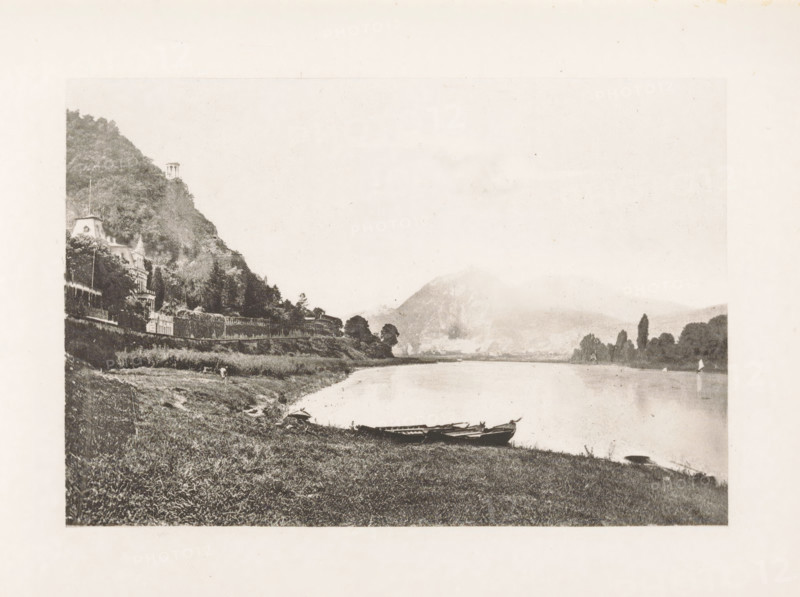 Street in Remagen, page, photomechanical print, title: 'Street in Remagen.', Remagen, c. 1889 - in or before 1899, Rhineland-Palatinate, paper, photoaquatint, height 132 mm x width 83 mm, street, A wide riverside scene in a faded sepia tone showing a grassy bank in the foreground with small rowboats pulled up at the water’s edge, a calm river stretching toward distant hazy hills and a pale sky, a wooded hillside on the left with ornate buildings partially visible among the trees and a small tower near the hilltop, a line of trees along the opposite shore and faint shapes of structures at the base of the distant hill.