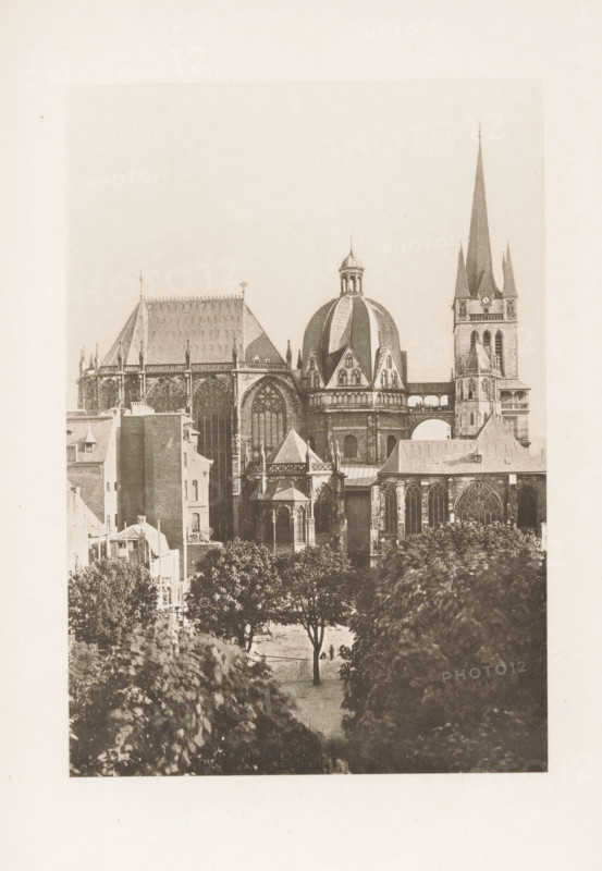 Exterior of Cologne Cathedral, page, photomechanical print, title: 'Cathedral of Cologne.', Cologne, c. 1889 - in or before 1899, North Rhine-Westphalia, paper, photoaquatint, height 124 mm x width 88 mm, church (exterior), Cologne Cathedral, A sepia-toned photograph of a large Gothic-style cathedral complex seen from a distance, showing a tall pointed spire flanked by smaller spires, a prominent domed roof with ribbed panels, large ornate arched windows with tracery, flying buttresses and decorative pinnacles, adjoining stone buildings with steep pitched roofs and smaller towers, a covered walkway or arch connecting sections, and trees and leafy foliage in the foreground partially obscuring the lower parts of the buildings.