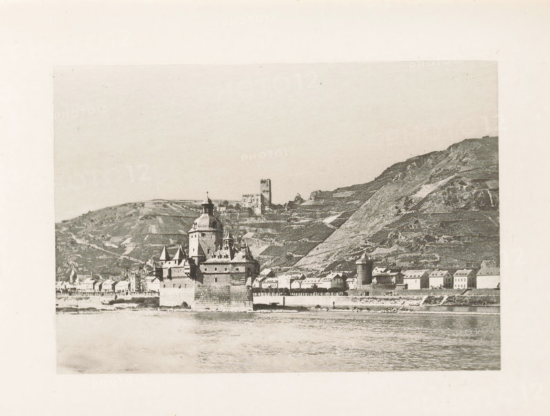View of Kaub and Burg Pfalzgravenstein, page, photomechanical print, title: 'Caub and the Pfalz.', Rhine, c. 1889 - in or before 1899, paper, photoaquatint, height 83 mm x width 120 mm, city-view in general, 'veduta', timber framing, Rhineland-Palatinate, Kaub, Pfaffenporte **, A sepia-toned photograph showing a riverside town with a waterfront wall and a large stone building featuring turrets and a central dome rising above smaller pitched roofs; behind the town, terraced hills slope upward with a solitary square tower on a ridge and scattered vineyard-like patterns on the hillside; the river occupies the foreground with rippled water reflecting the shoreline, and the scene is framed by a wide pale border.