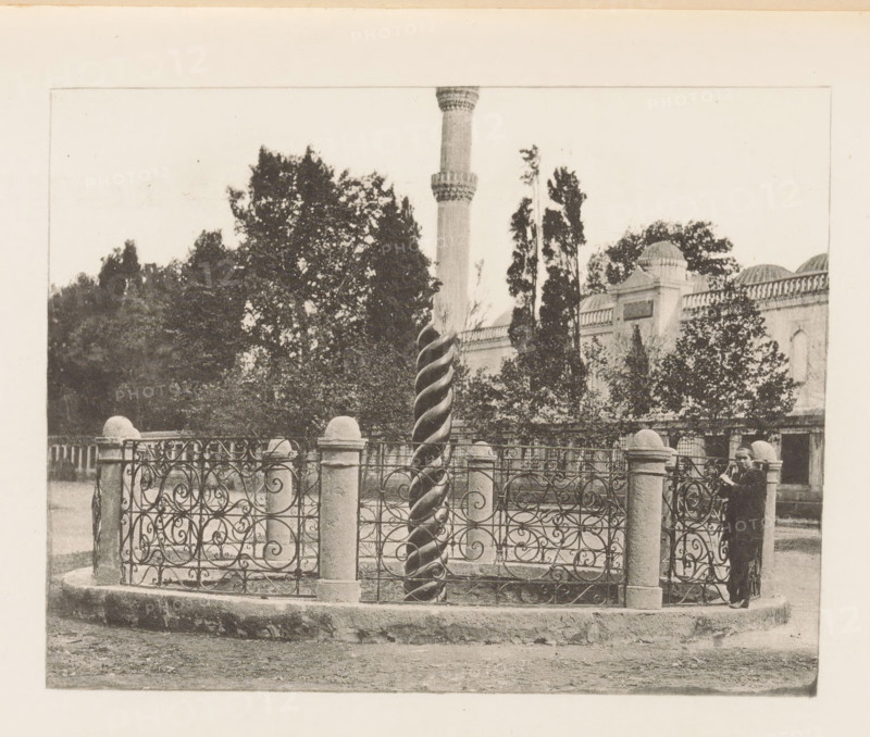 Serpentine Column in Constantinople, page, photomechanical print, title: 'Serpentine Column in the Hippodrome.', Constantinople, c. 1885 - in or before 1895, Ottoman Empire, paper, photoaquatint, height 94 mm x width 121 mm, column, pillar, architecture, Istanbul, Serpent Column, A black and white photograph shows an ornate wrought iron fence with swirling patterns supported by stone posts topped with rounded caps surrounding a tall twisted column. Behind the fence there are trees and a building with domed roof elements and arched windows, and a slender minaret rises above the foliage. A person stands near the fence on the right side, wearing a dark coat and hat and holding an object close to their face. The ground in front of the fenced area appears unpaved and textured.