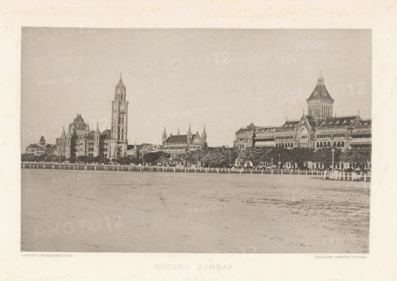 View of buildings in Mumbai, Maharashtra, India, page, photomechanical print, title: 'MODERN BOMBAY', signature, `: 'SAMPSON LOW MARSTON & CO. LTD, HÉLIOG & IMP LEMERCIER & CIE. PARIS', Mumbai, cliché maker: Lemercier & Cie., Paris, c. 1883 - in or before 1893, photographer, paper, photoaquatint, height 115 mm x width 177 mm, palace, façade (of house or building), A wide sepia-toned photograph shows a long row of ornate stone buildings with Gothic architectural features including pointed arches, steeply pitched roofs, towers, and a tall clock tower rising above the others, set behind a line of trees and a fence along a broad open foreground.