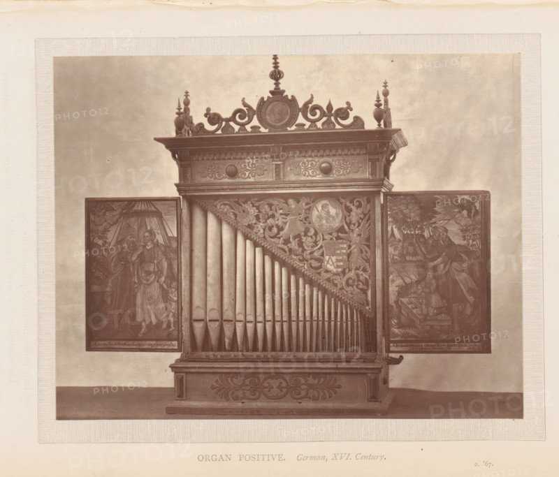 Organ of the South Kensington Museum in London, page, photomechanical print, inscription: 'Organ positive. German, XVI. Century.', London, c. 1869 - in or before 1874, paper, Woodburytype, height 116 mm x width 147 mm, organ, A photographic image of a wooden pipe organ with a decorative top featuring carved scrollwork and finials, a central circular medallion, a row of vertical organ pipes arranged in a triangular pattern, ornate carved panels with floral and figurative designs surrounding the pipes, and two hinged side panels displaying painted scenes of people and drapery, all set against a plain background and mounted on a carved base.