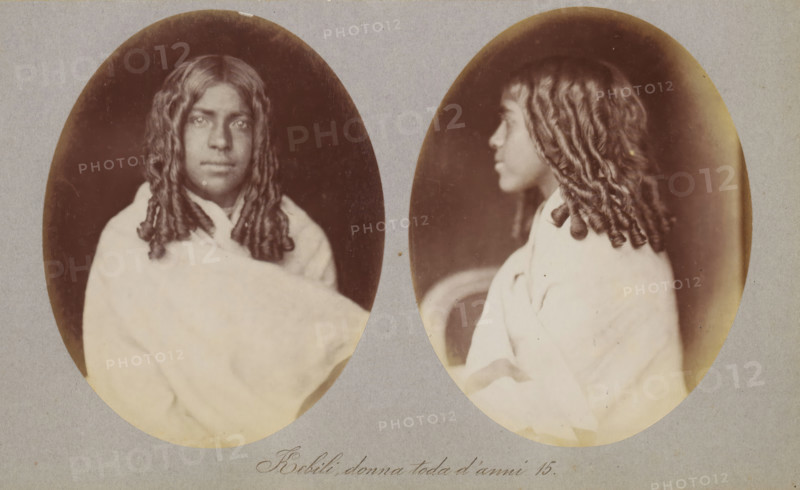 Kebili, donna toda d'anni 15, Two portraits of Kebili of the Toda tribe, photograph, page, photographer: Paolo Mantegazza, India, c. 1881 - in or before 1886, Paolo Mantegazza, photographer, paper, albumen print, height 151 mm x width 241 mm, adolescent, young woman, maiden (sideview, profile), maiden (front view), A sepia-toned double portrait showing the same young person in two oval frames side by side, one a front-facing view and the other a profile. The person has long, tightly curled or twisted hair falling past the shoulders and a neutral facial expression in the front view while the profile shows a slightly turned head. They are wrapped in a light-colored draped garment that covers the shoulders and chest. The ovals sit on a plain, grayish mount with a small area of handwritten text visible below the images. The background inside the ovals is dark, creating contrast with the subject and the light garment.