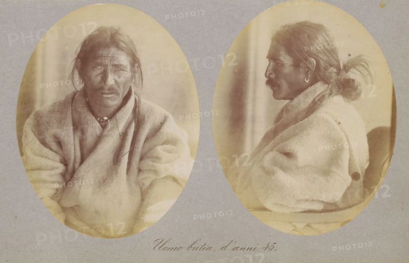 Two portraits of an unknown man, Uomo butia, d'anni 45, photograph, page, photographer: Paolo Mantegazza, Bhutan, c. 1881 - in or before 1886, Paolo Mantegazza, photographer, paper, albumen print, height 151 mm x width 241 mm, adult man (side view, profile), adult man (front view), Two oval sepia photographs mounted side by side on a gray background show the same middle-aged man from different angles; the left oval is a frontal view and the right oval is a profile view. He has long dark hair pulled back into a low tied style with a loose strand visible, a prominent mustache, and a weathered face with deep lines. He wears a thick, light-colored blanket or coat draped over his shoulders and a beaded necklace at the collar. In the profile image he has a small hoop earring in his visible ear and his hair is gathered into a knot at the back with loose wisps extending outward. The portraits have a soft, slightly faded tonal quality and the oval frames show a subtle uneven vignette around eac...
