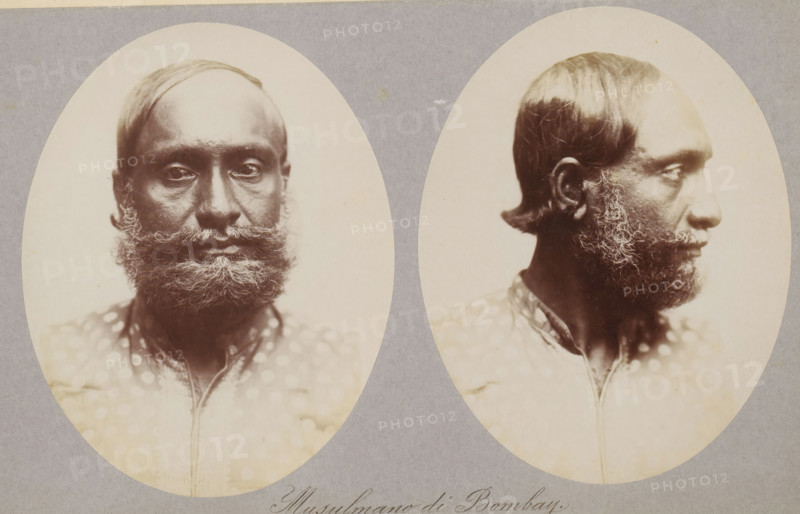Two portraits of an unknown man from Mumbai, Musulmano di Bombay, photograph, page, photographer: Paolo Mantegazza, Mumbai, c. 1881 - in or before 1886, Maharashtra, paper, albumen print, height 151 mm x width 241 mm, adult man (side view, profile), Two oval sepia-toned portrait photographs of the same man placed side by side on a gray background, one showing his face from the front and the other in profile. The man has short, dark hair with a receding hairline and a full, curly beard and mustache with lighter streaks. His skin appears dark and textured, and his eyes look straight ahead in the frontal image and to the side in the profile. He wears a patterned garment with a collar visible at the neckline. The photographs have soft lighting and show subtle variations in tone and detail against the plain background.