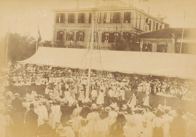 May-pole dance in Shanghai for the Diamond Jubilee of Victoria of the United Kingdom, The May-pole dance, photograph, page, photographer: WR Kahler (possibly), Shanghai, 1897, paper, albumen print, height 106 mm x width 151 mm, group dancing, commemoration, jubilee, festivities (wreath, garland, festive activities), A faded sepia photograph showing a large outdoor gathering in front of a multi-story building with shuttered windows and a covered awning; a tall pole with many ribbons extends upward from the center of the crowd where people in period clothing form a circle, while rows of spectators wearing hats fill the foreground and the area under the awning.