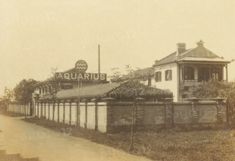 The Aquarius Company, Aquaris drinking water factory on Seward Road in Shanghai, photograph, page, Shanghai, c. 1884 - in or before 1894, photographer, paper, gelatin silver print, developed gelatin silver print, height 97 mm x width 140 mm, trade-company, A sepia photograph shows a street scene framed by a decorative border, featuring a long low wall with evenly spaced vertical sections and a tiled roof behind it, trees and shrubs scattered along the property, and a two-story building with a covered balcony, shuttered windows, and a hipped roof with a small roof ornament; a circular sign with a patterned emblem sits above the roofline near a taller vertical pole, and a paved road runs alongside the wall.