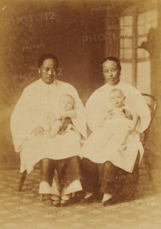 Portrait of two unknown Chinese nannies with babies on their laps, Chinese Amahs, photograph, page, China, c. 1884 - in or before 1894, photographer, paper, albumen print, height 144 mm x width 103 mm, baby-sitter, A faded sepia photograph bordered by decorative lines shows two seated women wearing light-colored tops and darker skirts, each holding an infant in white clothing on their laps; the women have similar hairstyles pulled back and neutral facial expressions, and the background includes a window with panes and a patterned floor beneath their feet.