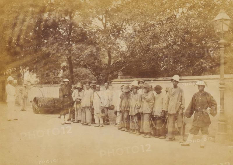 Group of Chinese people chained together under the supervision of guards, The chain-gang, photograph, page, China, c. 1884 - in or before 1894, photographer, paper, albumen print, height 102 mm x width 144 mm, workhouse, penal camp, penitentiary, chain gang, A sepia-toned photograph mounted on a decorative bordered page shows a group of people standing in a line along a street in front of a low wall with trees behind it; most individuals wear loose clothing and hats, and several are connected by chains; two men in lighter uniforms stand near a large cylindrical object on the left, while a man in a darker uniform stands by a tall streetlamp on the right; the scene includes a paved ground and dappled light from the foliage above.