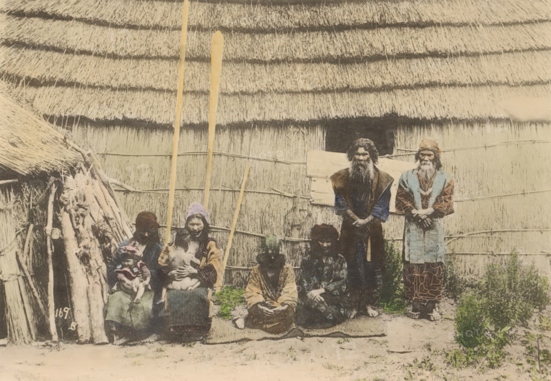Ainues, the Native of Hokkaido, Japan, Portrait of unknown people of the Aino in front of a house in Japan, page, photomechanical print, number: '169.B', maker: Kozaburo Tamamura (possibly), c. 1895 - c. 1905, paper, collotype, photoprint, hand colored, height 105 mm x width 151 mm, human types, peoples and nationalities, A faded photograph shows a group of people posed in front of a large thatched wall and roof made of layered straw. Several tall wooden poles lean upright against the wall. Two older men stand at the back with long beards and layered clothing, one wearing a patterned garment and the other wearing a vest over darker sleeves. In front of them three seated adults wear varied patterned and textured clothing, one holding a small child and another holding an infant; one seated person has a head covering. To the left a simple structure of sticks and thatch is visible with bundled wood and rough supports. The ground in front of the group is bare earth with patches of low veget...